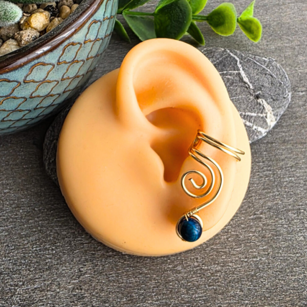 Handgefertigter, hellgold vergoldeter Ear Cuff Vortex von MaMari Art mit einem echten blauen Tigerauge, getragen an einem realistischen Silikonohr zur Demonstration des Sitzes ohne Piercing.