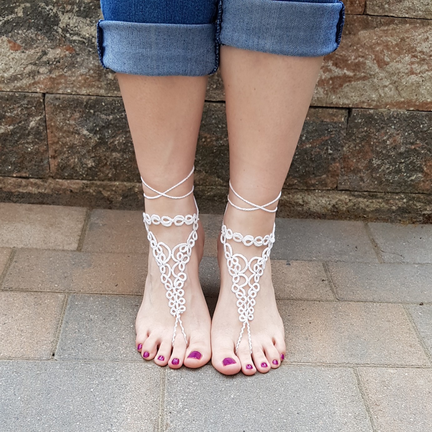 Eine Frau trägt handgefertigte weiße Barfuß-Sandalen aus feiner Occhi-Knotentechnik. Die filigranen Ornamente verlaufen elegant vom Knöchel bis zu den Zehen und werden mit zarten Bändern am Bein gebunden. Die Füße stehen auf einem gepflasterten Boden, im Hintergrund ist eine rustikale Steinmauer zu sehen. Die Zehennägel sind in einem dunklen Beerenton lackiert, die Jeans lässig hochgekrempelt.