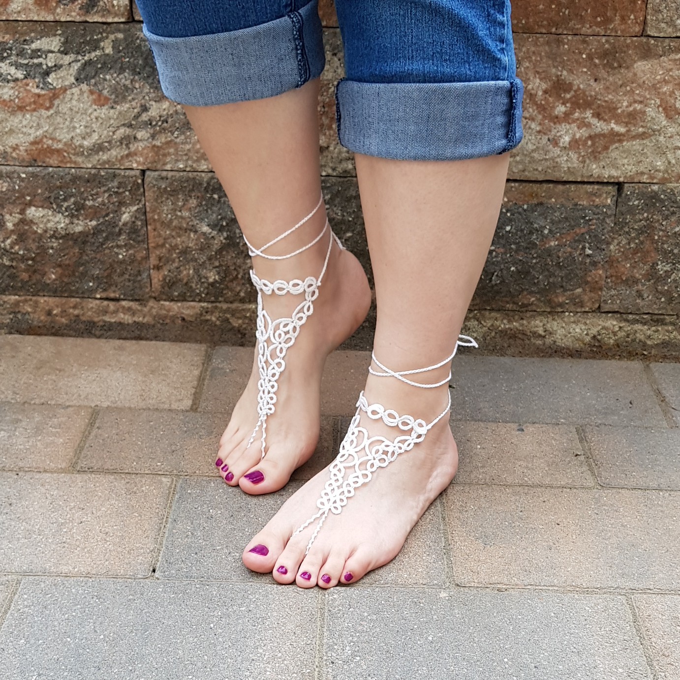 Eine Frau trägt handgefertigte weiße Barfuß-Sandalen aus feiner Occhi-Knotentechnik. Die filigranen Ornamente verlaufen elegant vom Knöchel bis zu den Zehen und werden mit zarten Bändern am Bein gebunden. Die Füße stehen auf einem gepflasterten Boden, im Hintergrund ist eine rustikale Steinmauer zu sehen. Die Zehennägel sind in einem dunklen Beerenton lackiert, die Jeans lässig hochgekrempelt.
