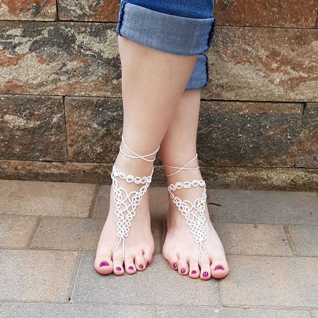 Eine Frau trägt handgefertigte weiße Barfuß-Sandalen aus feiner Occhi-Knotentechnik. Die filigranen Ornamente verlaufen elegant vom Knöchel bis zu den Zehen und werden mit zarten Bändern am Bein gebunden. Die Füße stehen auf einem gepflasterten Boden, im Hintergrund ist eine rustikale Steinmauer zu sehen. Die Zehennägel sind in einem dunklen Beerenton lackiert, die Jeans lässig hochgekrempelt.