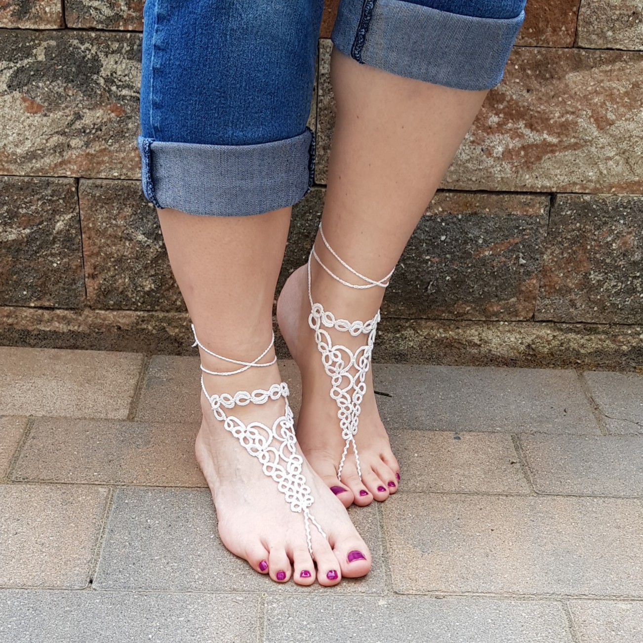 Eine Frau trägt handgefertigte weiße Barfuß-Sandalen aus feiner Occhi-Knotentechnik. Die filigranen Ornamente verlaufen elegant vom Knöchel bis zu den Zehen und werden mit zarten Bändern am Bein gebunden. Die Füße stehen auf einem gepflasterten Boden, im Hintergrund ist eine rustikale Steinmauer zu sehen. Die Zehennägel sind in einem dunklen Beerenton lackiert, die Jeans lässig hochgekrempelt.