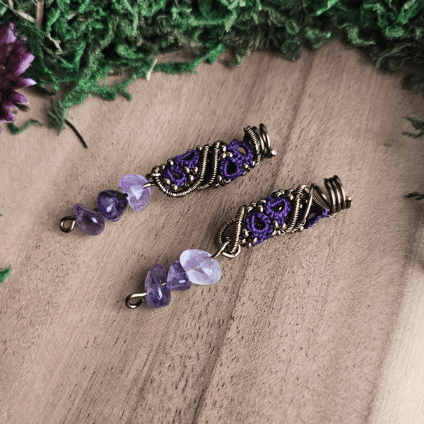 Ein Paar handgefertigter Dreadspiralen von MaMari Art liegt auf einer Holzoberfläche. Die Spiralen bestehen aus bronzefarbenem Draht, der kunstvoll um ein filigranes Occhi-Muster in leuchtendem Violett gewickelt ist. Kleine bronzefarbene Perlen sind in das Muster eingearbeitet und setzen feine Akzente. Am unteren Ende der Spiralen hängen jeweils drei polierte Splitter aus Amethyst in verschiedenen Lilaschattierungen. Der Hintergrund ist mit grünem Moos und getrockneten Pflanzen dekorativ gestaltet und verstärkt die naturverbundene Ausstrahlung des Schmucks.
