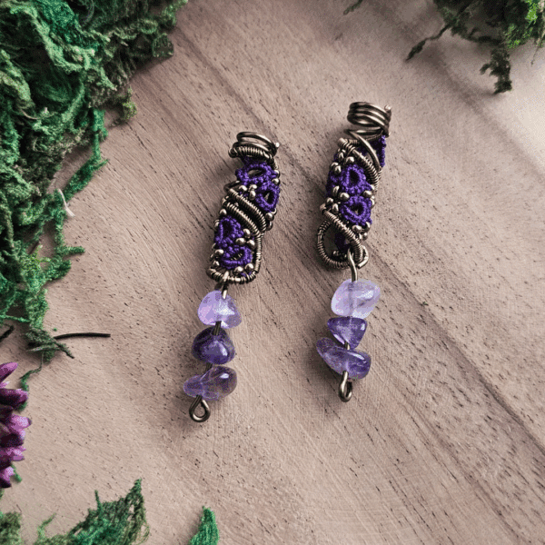Ein Paar handgefertigter Dreadspiralen von MaMari Art liegt auf einer Holzoberfläche. Die Spiralen bestehen aus bronzefarbenem Draht, der kunstvoll um ein filigranes Occhi-Muster in leuchtendem Violett gewickelt ist. Kleine bronzefarbene Perlen sind in das Muster eingearbeitet und setzen feine Akzente. Am unteren Ende der Spiralen hängen jeweils drei polierte Splitter aus Amethyst in verschiedenen Lilaschattierungen. Der Hintergrund ist mit grünem Moos und getrockneten Pflanzen dekorativ gestaltet und verstärkt die naturverbundene Ausstrahlung des Schmucks.
