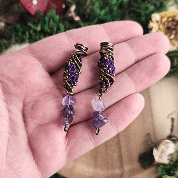 Eine Hand hält ein Paar handgefertigter Dreadspiralen von MaMari Art. Die Spiralen bestehen aus bronzefarbenem Draht, der kunstvoll um ein detailreiches Occhi-Muster in kräftigem Violett gewickelt ist. Kleine bronzefarbene Perlen betonen die Struktur und setzen glänzende Akzente. Am unteren Ende hängen jeweils drei polierte Amethystsplitter in unterschiedlichen Lilatönen. Der natürliche Hintergrund mit Moos, Holz und getrockneten Pflanzen unterstreicht den erdigen und fantasievollen Charakter der Schmuckstücke.