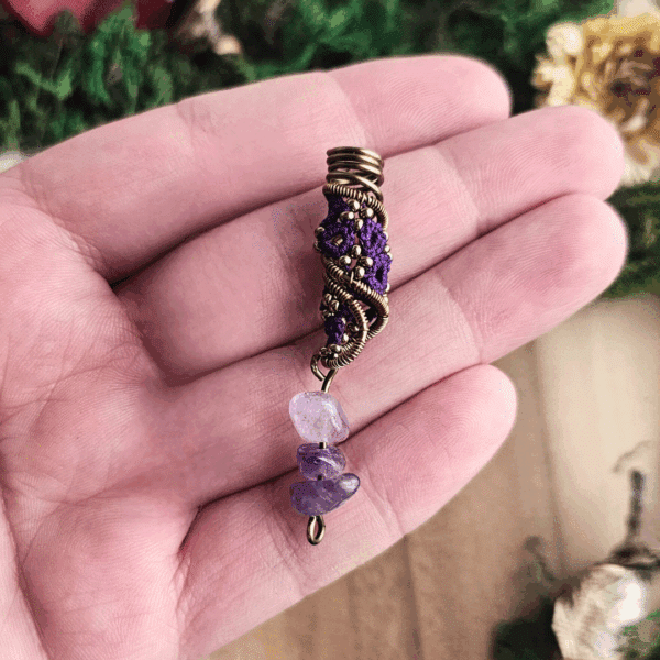 Eine Hand hält eine handgefertigte Dreadspirale von MaMari Art. Die Spirale besteht aus bronzefarbenem Draht, kunstvoll um ein Occhi-Muster in kräftigem Violett gewickelt. Kleine bronzefarbene Perlen sind in das Muster eingearbeitet und verleihen dem Design zusätzliche Tiefe und Glanz. Am unteren Ende hängen drei polierte Splitter aus Amethyst in verschiedenen Lilatönen, die das Farbschema harmonisch ergänzen. Der Hintergrund mit Moos und getrockneten Pflanzen unterstreicht den naturverbundenen Charakter des Schmucks.