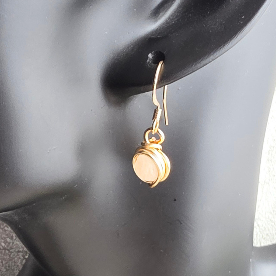 Goldfarbener handgefertigter Ohrring mit eingefasster weiß schimmernder Perle an einer schwarzen Schmuckbüste. Der kleine Anhänger ist mit Draht umwickelt und hängt an einem filigranen Ohrhaken.