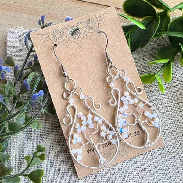 Eine schräge Nahaufnahme der Wirework-Ohrringe Lebensbaum auf einer Schmuckkarte; betont die ovale Tropfenform, die AB-Glasperlen und die hochwertige Versilberung