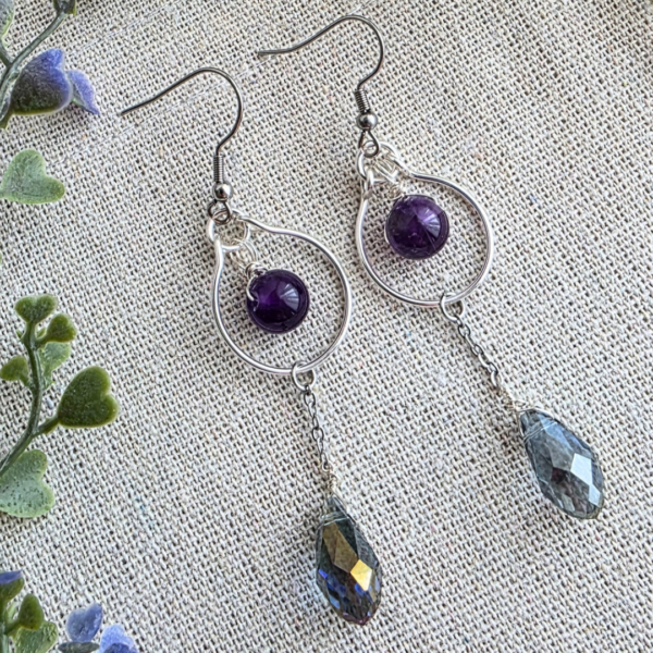 Seitliche Nahaufnahme der MaMari Art Ohrringe Elfenträne auf Leinen; betont die facettierten Glastropfen, die Amethystperlen und die zarte Edelstahlkette.