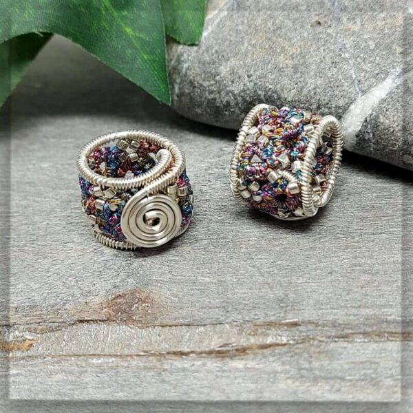 Zwei handgefertigte Dreadperlen aus versilbertem Kupferdraht mit buntem Polyestergarn und silbernen Glasperlen. Eine der Perlen zeigt ein spiralförmiges Design, die andere ist detailreich verziert. Sie liegen auf einer grauen Oberfläche neben einem Stein und einem grünen Blatt.