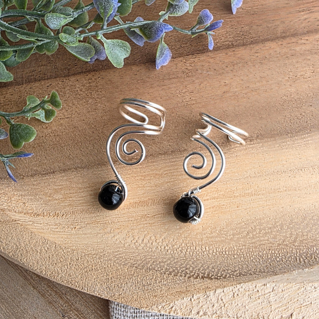 Zwei silberfarbene Ear Cuffs Vortex mit Spiralen und schwarzen Onyxperlen auf natürlichem Holzhintergrund.