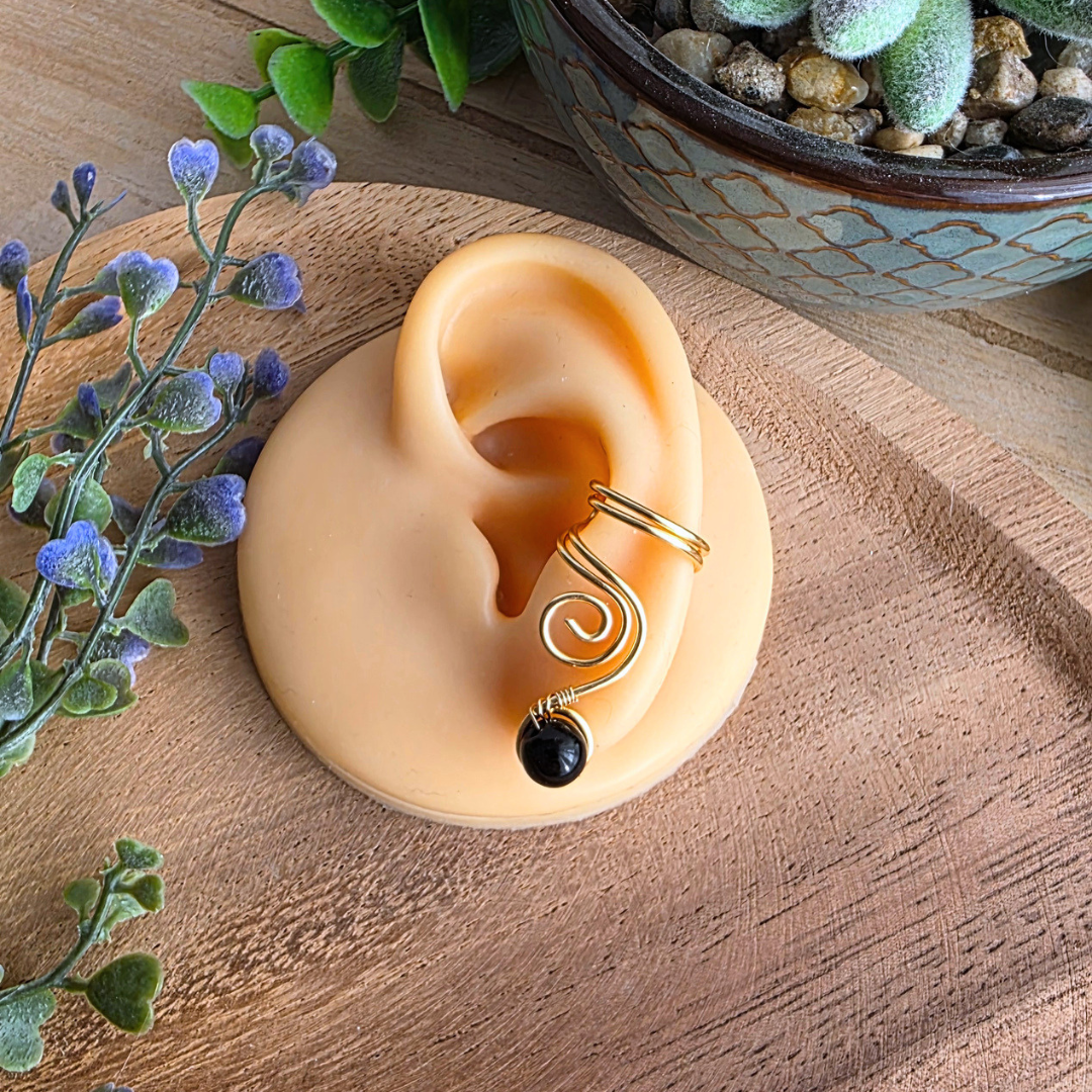 Goldfarbener Ear Cuff Vortex mit schwarzer Onyxperle an einem Silikonohr zur Darstellung der Trageweise.