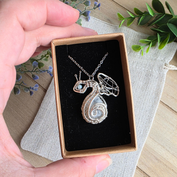 Detailaufnahme der Halskette „Drache Mondhüter“ in einer kleinen Schmuckschachtel, gehalten in einer Hand. Der Drachenanhänger aus silberfarbenem Drahtkunst-Design umschließt einen schimmernden Mondstein und ist mit einem hellblauen Amazonit verziert.