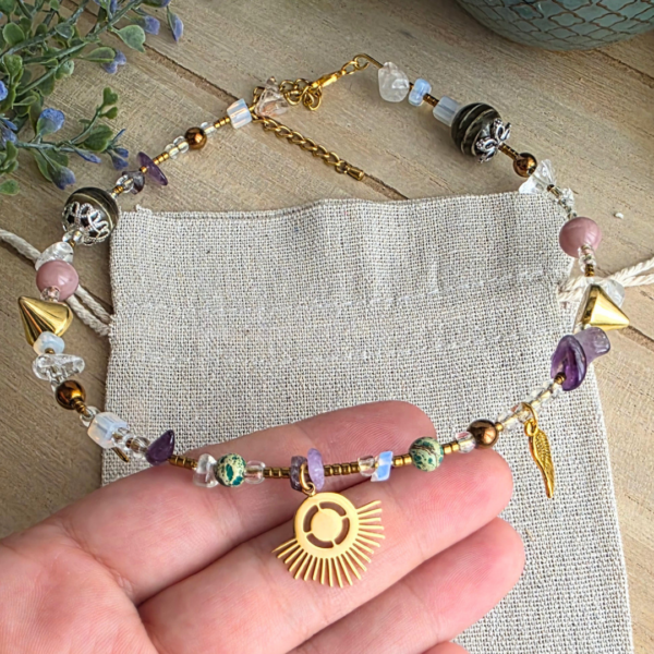 Nahaufnahme des goldfarbenen Sonnenanhängers an einem Halsreif mit Amethystsplittern, grünen Steinperlen, Glasperlen und einem kleinen Flügelanhänger
