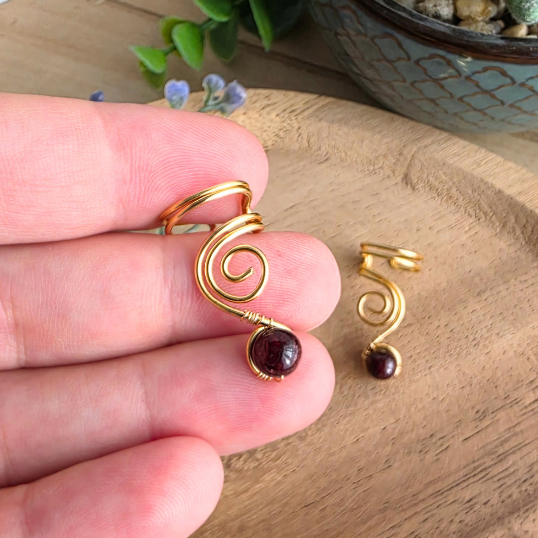 Ear Cuff Vortex Goldfarben mit Granat in der Hand gehalten vor einem Holzhintergrund