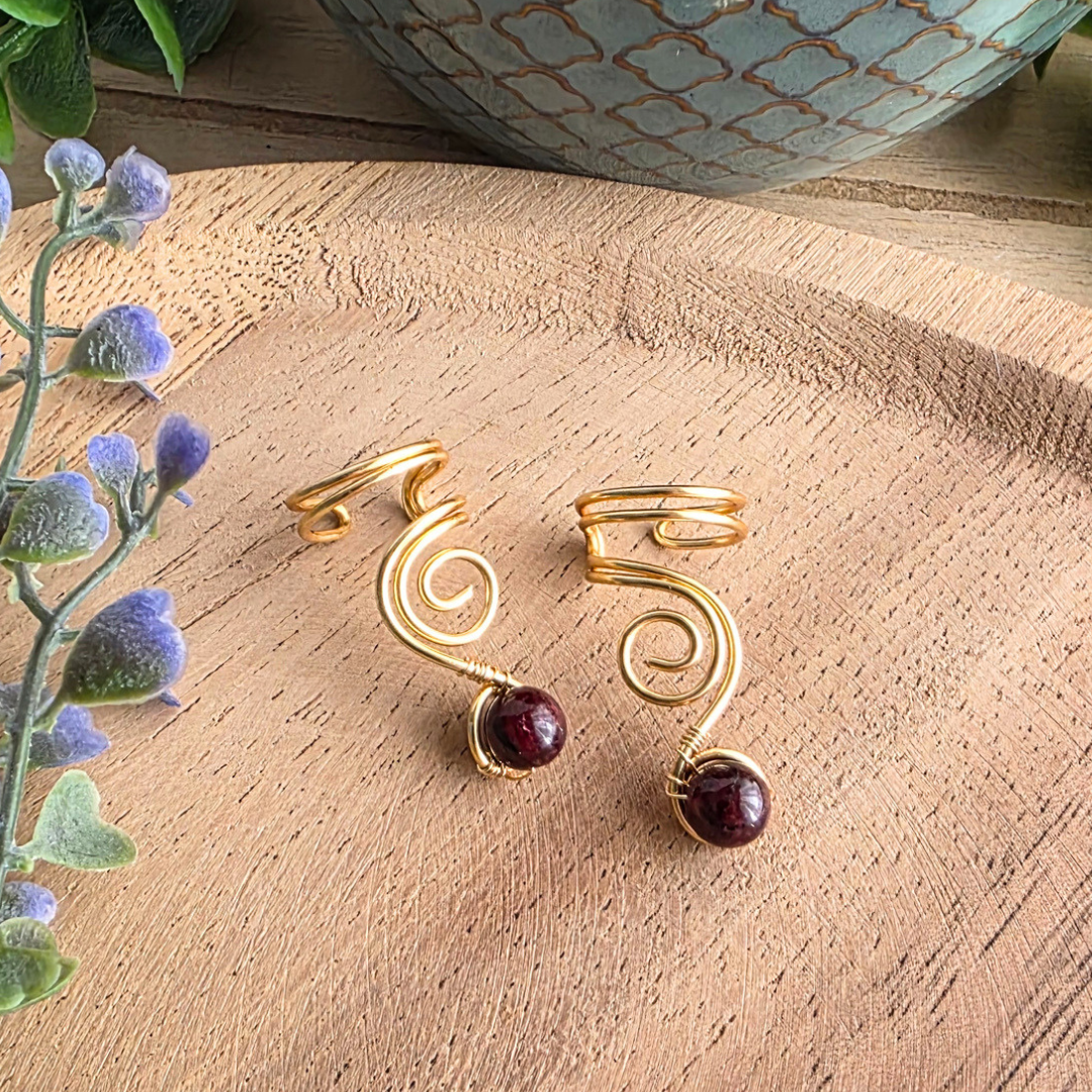 1 Paar Ear Cuff Vortex Goldfarben mit Granat auf einem Holzhintergrund und lila Blumen