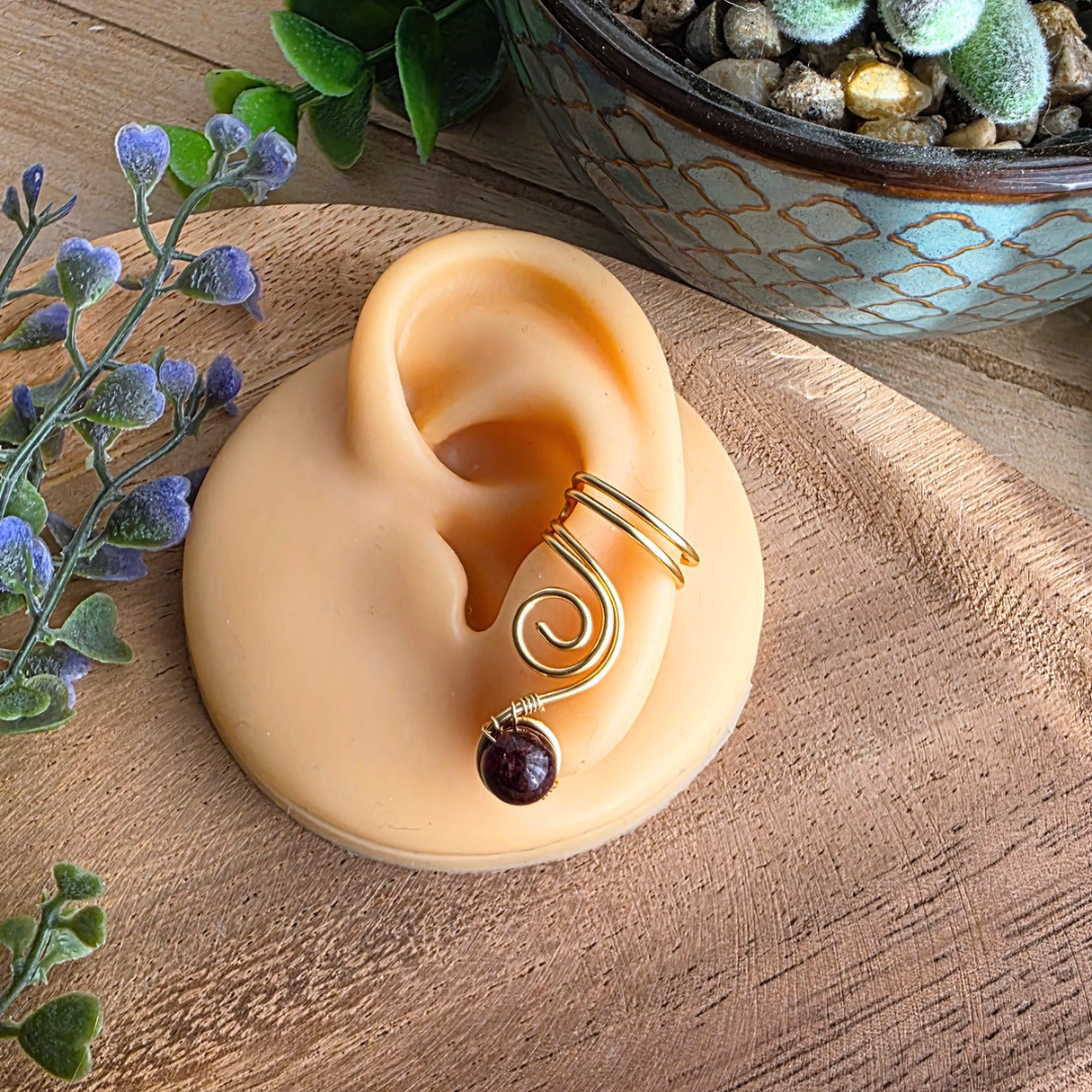 Ear Cuff Vortex Goldfarben Granat auf einem Silikonohr auf Holzhintergrund