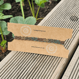 Ein handgefertigtes Fußkettchen von MaMari Art ist auf einer braunen Kraftpapier-Karte befestigt, die mit weißen Mandala-Elementen bedruckt ist. In der Mitte steht „handgefertigt von“ und darunter die Website www.mamari-art.de. Das Fußkettchen besteht aus Occhi-Spitze in verschiedenen Naturtönen wie Braun, Grün, Blau und Senfgelb und zeigt ein detailliertes, florales Muster. Die Präsentation erfolgt im Freien auf einer Holzbank, im Hintergrund wachsen grüne Erdbeerpflanzen in einem Beet.