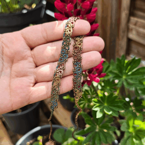 Eine Hand hält ein handgefertigtes Fußkettchen von MaMari Art in die Sonne. Das Schmuckstück besteht aus fein gearbeiteter Occhi-Spitze in einem harmonischen Farbverlauf aus Braun, Grün, Blau und Senfgelb. Kleine goldfarbene Perlen akzentuieren das filigrane Muster. Im Hintergrund blühen pinkfarbene Lupinen, umgeben von grünen Blättern und Pflanztöpfen auf einer Holzterrasse. Die Szene vermittelt eine sommerliche, natürliche Stimmung.