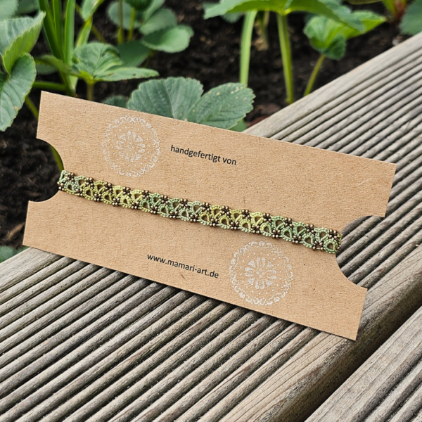 Grünes handgefertigtes Fußkettchen mit antikbronzenen Glasperlen, liebevoll auf einer Kraftpapier-Schmuckkarte präsentiert. Die Karte liegt auf einer sonnenbeschienenen Holzterrasse vor einem Beet mit Erdbeerpflanzen. Auf der Karte stehen „handgefertigt von“ und „www.mamari-art.de“