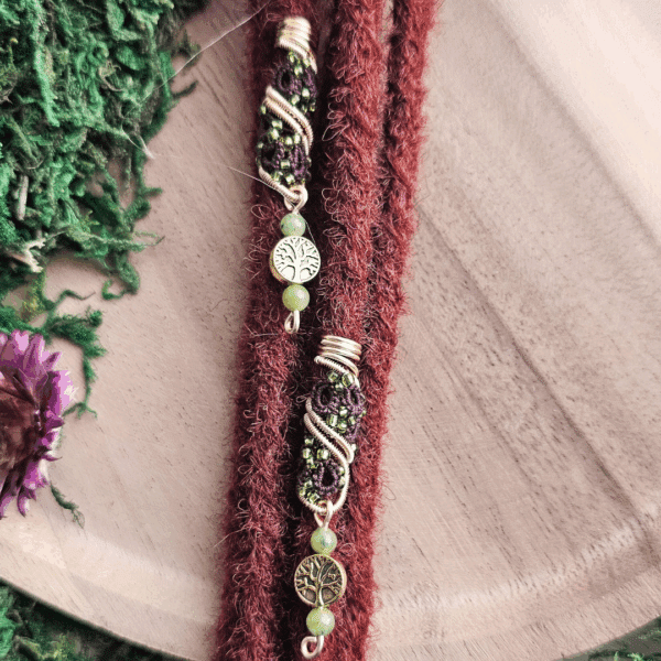 Eine handgefertigte Dreadspirale von MaMari Art ist in zwei rötlich-braune Dreadlocks eingearbeitet. Die Spirale besteht aus goldfarbenem Draht, der kunstvoll gewickelt ist und eine filigrane Makramee-Verzierung in dunklem Violett und Grün umschließt. Kleine grüne Perlen und metallene Anhänger mit Lebensbaum-Motiv ergänzen das Design. Die Dreadspirale wird auf einer Holzunterlage präsentiert, umgeben von Moos und Trockenblumen, was die naturverbundene Ästhetik des Schmucks unterstreicht.