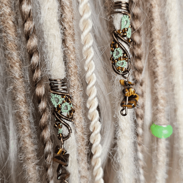 Zwei handgefertigte Dreadspiralen von MaMari Art sind in helle, naturfarbene Dreadlocks eingeflochten. Die Spiralen bestehen aus bronzefarbenem Draht, der kunstvoll um ein filigranes Occhi-Muster in Mintgrün gewunden ist. Orange-braune Glasperlen setzen glänzende Akzente entlang der Spirale. Am unteren Ende hängen kleine Splitter aus Tigerauge, die dem Schmuckstück eine erdige, naturverbundene Note verleihen. Die Dreadlocks im Hintergrund variieren in Creme-, Beige- und Brauntönen und unterstreichen den harmonischen Boho-Stil.