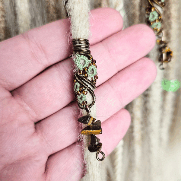 Eine handgefertigte Dreadspirale von MaMari Art ist in eine helle, cremefarbene Dreadlock eingearbeitet und wird von einer Hand gehalten. Die Spirale besteht aus bronzefarbenem Draht, der kunstvoll um ein mintgrünes Occhi-Muster gewickelt ist. Orange-braune Glasperlen setzen schimmernde Akzente entlang des Designs. Am unteren Ende der Spirale hängen kleine, polierte Splitter aus Tigerauge, die dem Schmuckstück eine erdige und natürliche Ausstrahlung verleihen. Im Hintergrund sind weitere Dreadlocks in verschiedenen Beige- und Brauntönen zu sehen.