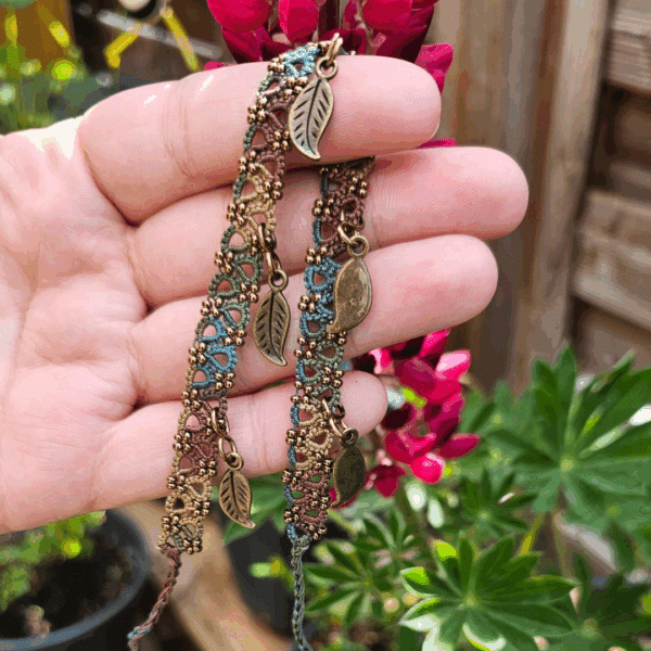 Eine Hand hält ein handgefertigtes Fußkettchen von MaMari Art mit kleinen bronzefarbenen Blattanhängern in die Sonne. Das Fußkettchen besteht aus fein geknoteter Occhi-Spitze in einem harmonischen Farbverlauf aus Braun, Grün, Blau und Senfgelb. Goldfarbene Perlen akzentuieren das detaillierte Muster. Die Blattanhänger sind gleichmäßig entlang des Fußkettchens verteilt und bewegen sich frei. Im Hintergrund blühen pinkfarbene Lupinen, umgeben von grünem Laub und Pflanztöpfen auf einer Holzterrasse – die Szene strahlt sommerliche Leichtigkeit und Naturverbundenheit aus.