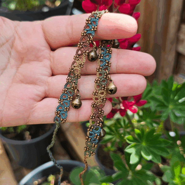 Eine Hand hält ein handgefertigtes Fußkettchen von MaMari Art in die Sonne. Das Kettchen besteht aus fein geknoteter Occhi-Spitze in einem natürlichen Farbverlauf aus Türkis, Braun, Grün, Senfgelb und Altrosa. Kleine bronzefarbene Glöckchen sind gleichmäßig entlang des Bandes angebracht und schwingen locker mit. Die Szene spielt auf einer Terrasse, im Hintergrund sind grüne Blätter, Blumentöpfe und pinkfarbene Lupinenblüten zu sehen – sie unterstreichen die sommerliche und naturverbundene Ästhetik des Schmuckstücks.