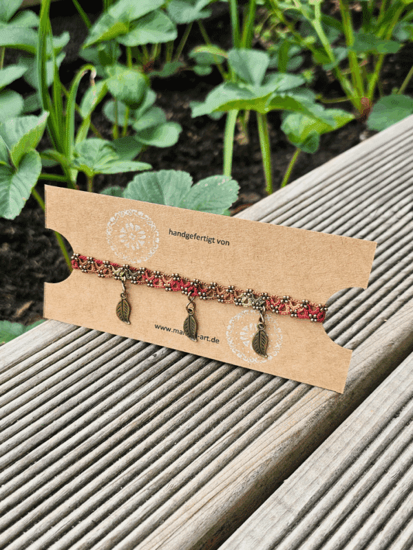 Handgefertigtes Fußkettchen auf einer Kraftpapierkarte mit dem Logo und der Website von MaMari Art. Das Fußkettchen besteht aus fein geknoteter Occhi-Spitze in warmen Herbstfarben – Terrakotta, Orange, Olive und Mocha – und ist mit antikbronzenen Blattanhängern verziert. Die Karte liegt auf einer Holzterrasse im Garten, im Hintergrund wachsen grüne Erdbeerpflanzen im Hochbeet.