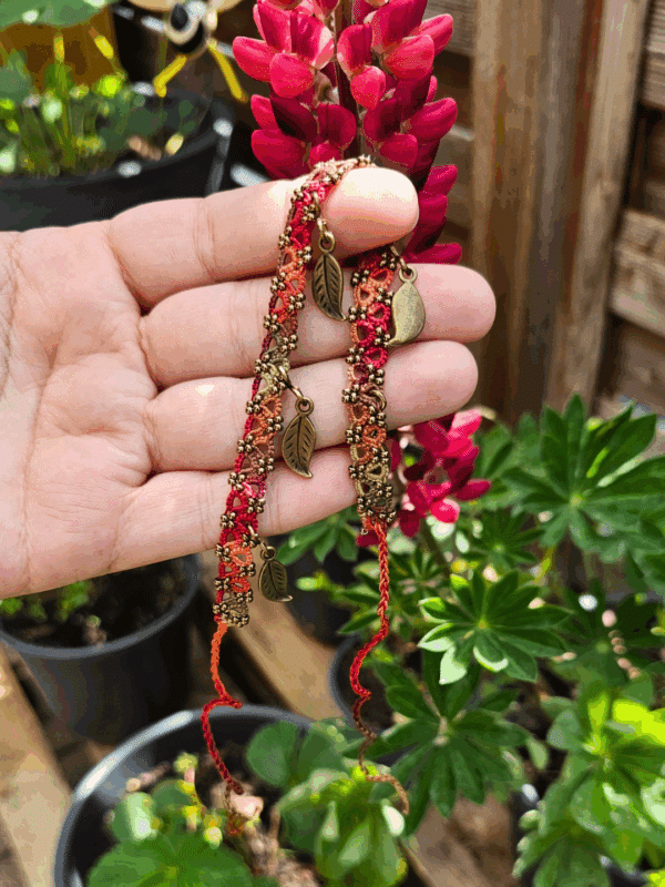 Fußkettchen aus Occhi-Spitze in herbstlichen Farben wie Orange, Rot, Moosgrün und Terrakotta, verziert mit kleinen antikbronzenen Blattanhängern. Das Schmuckstück wird von einer Hand vor einer pink blühenden Lupine in einem Garten gehalten. Im Hintergrund sind grüne Blätter und Pflanztöpfe zu sehen.