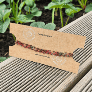 Ein handgefertigtes Fußkettchen von MaMari Art ist auf einer schlichten braunen Schmuckkarte mit weißem Mandala-Motiv und der Website-Adresse präsentiert. Das Kettchen zeigt ein lebendiges Farbverlaufsgarn in herbstlichen Tönen wie Mocha-Braun, Terrakotta, Orange, Goldgelb und Olivgrün. Kleine antikbronze Glasperlen betonen die kunstvoll geknotete Occhi-Spitze. Die Karte liegt auf einer hölzernen Terrasse, im Hintergrund wachsen grüne Erdbeerpflanzen, was eine natürliche und saisonale Stimmung vermittelt.