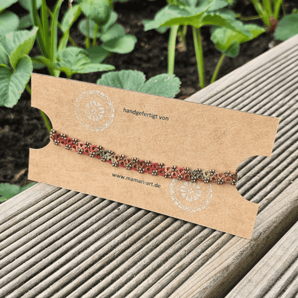 Ein handgefertigtes Fußkettchen von MaMari Art ist auf einer schlichten braunen Schmuckkarte mit weißem Mandala-Motiv und der Website-Adresse präsentiert. Das Kettchen zeigt ein lebendiges Farbverlaufsgarn in herbstlichen Tönen wie Mocha-Braun, Terrakotta, Orange, Goldgelb und Olivgrün. Kleine antikbronze Glasperlen betonen die kunstvoll geknotete Occhi-Spitze. Die Karte liegt auf einer hölzernen Terrasse, im Hintergrund wachsen grüne Erdbeerpflanzen, was eine natürliche und saisonale Stimmung vermittelt.