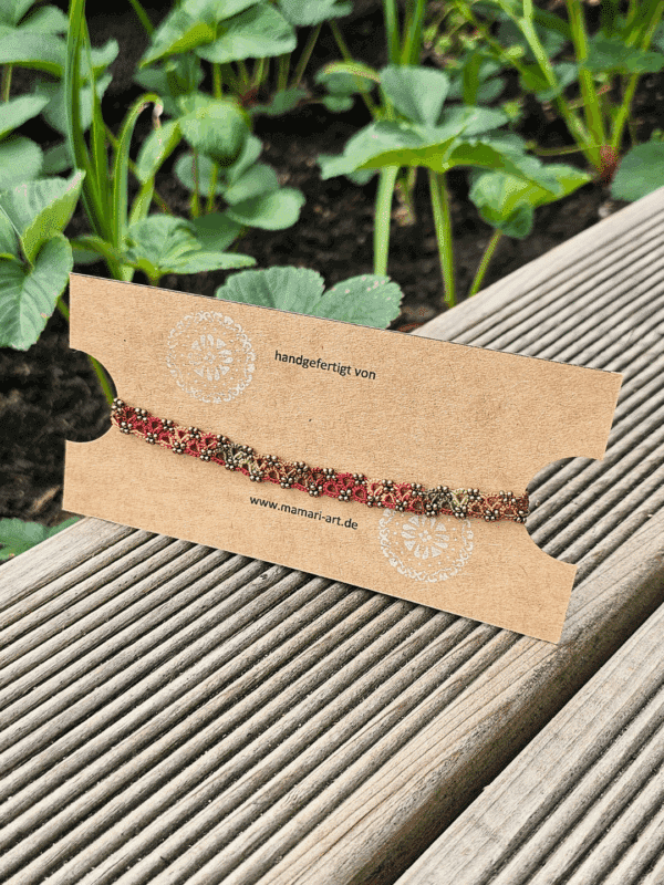 Ein handgefertigtes Fußkettchen von MaMari Art ist auf einer schlichten braunen Schmuckkarte mit weißem Mandala-Motiv und der Website-Adresse präsentiert. Das Kettchen zeigt ein lebendiges Farbverlaufsgarn in herbstlichen Tönen wie Mocha-Braun, Terrakotta, Orange, Goldgelb und Olivgrün. Kleine antikbronze Glasperlen betonen die kunstvoll geknotete Occhi-Spitze. Die Karte liegt auf einer hölzernen Terrasse, im Hintergrund wachsen grüne Erdbeerpflanzen, was eine natürliche und saisonale Stimmung vermittelt.