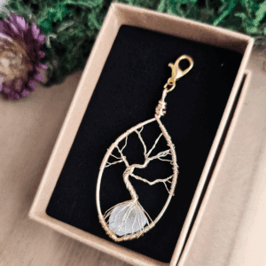 In einer offenen, naturfarbenen Schmuckbox mit schwarzem Inlay liegt ein filigraner Lebensbaum-Anhänger aus goldfarbenem Draht. Der Baum ist kunstvoll in einen tropfenförmigen Rahmen eingearbeitet, mit einem klaren Bergkristall in den Wurzeln. Der Anhänger ist mit einem goldfarbenen Karabinerhaken versehen. Im Hintergrund liegen grüne Dekomaterialien und eine getrocknete Blüte.