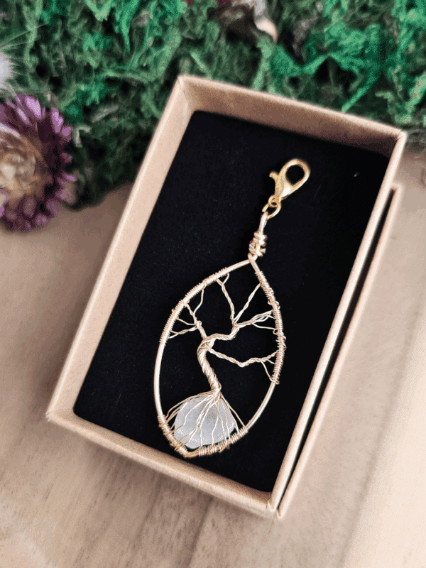 In einer offenen, naturfarbenen Schmuckbox mit schwarzem Inlay liegt ein filigraner Lebensbaum-Anhänger aus goldfarbenem Draht. Der Baum ist kunstvoll in einen tropfenförmigen Rahmen eingearbeitet, mit einem klaren Bergkristall in den Wurzeln. Der Anhänger ist mit einem goldfarbenen Karabinerhaken versehen. Im Hintergrund liegen grüne Dekomaterialien und eine getrocknete Blüte.