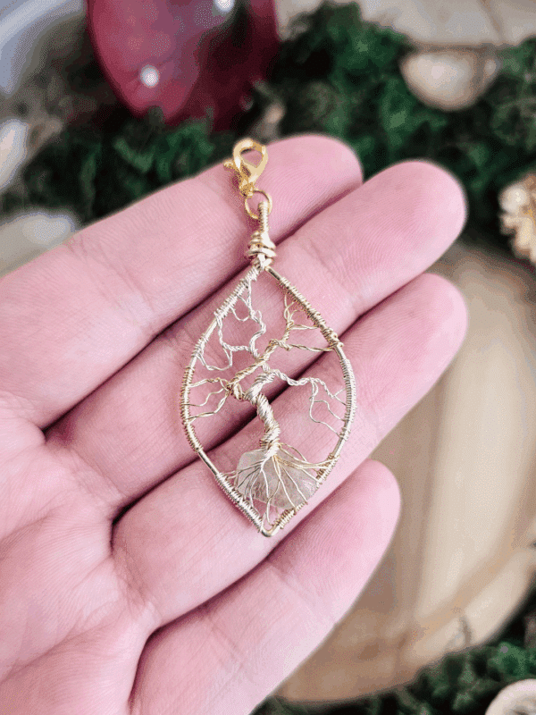 Ein handgefertigter Lebensbaum-Anhänger aus silber- und goldfarbenem Draht wird in einer offenen Hand präsentiert. Der tropfenförmige Rahmen umfasst einen Baum mit verdrehtem Stamm und fein gearbeiteten Ästen, dessen Wurzeln einen Bergkristallsplitter umschließen. Oben befindet sich ein goldfarbener Karabinerhaken. Im Hintergrund sind Moos, Holz und getrocknete Blumen unscharf zu erkennen.