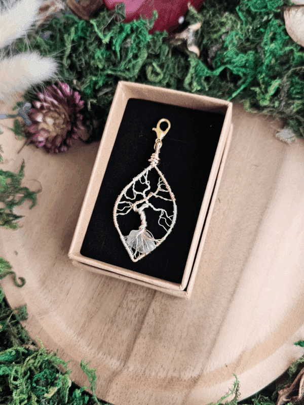Ein filigraner Lebensbaum-Anhänger aus silber- und goldfarbenem Draht liegt in einer schwarzen Schmuckeinlage in einer braunen Geschenkbox. Die Wurzeln umschlingen einen transparenten Bergkristallsplitter. Der Anhänger ist tropfenförmig gebogen und oben mit einem goldfarbenen Karabiner versehen. Um die Box herum sind Moos, getrocknete Blumen und ein Holztablett dekorativ arrangiert.
