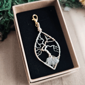Ein handgefertigter Lebensbaum-Anhänger aus silber- und goldfarbenem Draht liegt in einer Schmuckschachtel mit schwarzer Einlage. Die feinen Äste des kunstvoll gedrehten Drahtbaums erstrecken sich nach oben, während die Wurzeln einen transparenten Bergkristall umschließen. Der Anhänger ist tropfenförmig und mit einem goldfarbenen Karabinerhaken versehen. Die Box steht auf einer hellen Holzoberfläche, umgeben von grünem Dekomaterial.