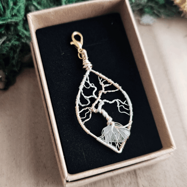 Ein handgefertigter Lebensbaum-Anhänger aus silber- und goldfarbenem Draht liegt in einer Schmuckschachtel mit schwarzer Einlage. Die feinen Äste des kunstvoll gedrehten Drahtbaums erstrecken sich nach oben, während die Wurzeln einen transparenten Bergkristall umschließen. Der Anhänger ist tropfenförmig und mit einem goldfarbenen Karabinerhaken versehen. Die Box steht auf einer hellen Holzoberfläche, umgeben von grünem Dekomaterial.