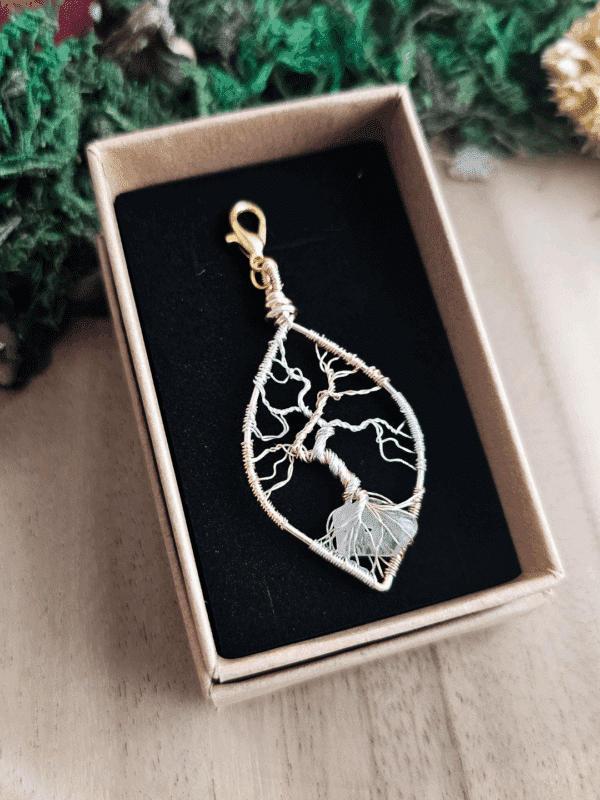 Ein handgefertigter Lebensbaum-Anhänger aus silber- und goldfarbenem Draht liegt in einer Schmuckschachtel mit schwarzer Einlage. Die feinen Äste des kunstvoll gedrehten Drahtbaums erstrecken sich nach oben, während die Wurzeln einen transparenten Bergkristall umschließen. Der Anhänger ist tropfenförmig und mit einem goldfarbenen Karabinerhaken versehen. Die Box steht auf einer hellen Holzoberfläche, umgeben von grünem Dekomaterial.