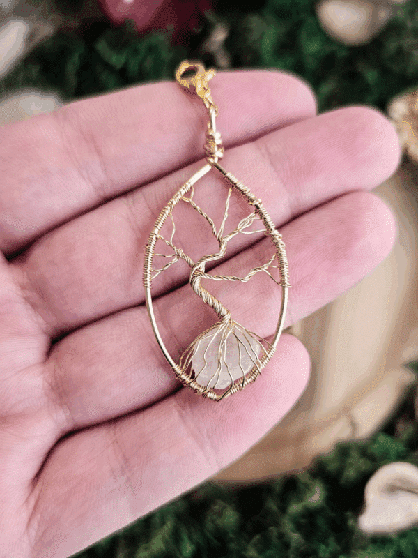 Ein handgefertigter Lebensbaum-Anhänger aus goldfarbenem Draht liegt auf einer geöffneten Handfläche. Der filigrane Baum ist in einen tropfenförmigen Rahmen eingearbeitet, seine Wurzeln umfassen einen klaren Bergkristall. Oben ist ein goldfarbener Karabinerhaken befestigt. Im Hintergrund ist unscharf natürliche Deko mit Moos und Trockenblumen zu erkennen.