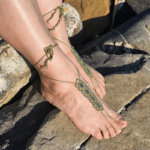 Zwei nackte Füße tragen handgefertigte Barfuß-Sandalen aus olivgrünem Garn mit filigranem Blumenmuster in hellblau und silber. Die Sandalen sind auf dem Spann geknotet und werden um die Knöchel gewickelt. Die Füße ruhen auf einem sonnenbeschienenen, unebenen Felsen, der natürliche Farben von Beige bis Dunkelgrau zeigt. Die Szene wirkt sommerlich und naturverbunden.