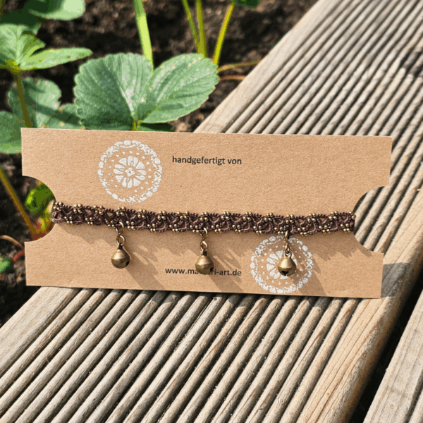 Handgefertigtes braunes Fußkettchen mit kleinen Glöckchen, liebevoll auf einer Kraftpapier-Karte präsentiert. Die feine Knotentechnik ist mit winzigen Glasperlen in Bronze- und Goldtönen verziert. Im Hintergrund sieht man ein Hochbeet mit Erdbeerpflanzen, das Schmuckstück liegt auf einer Holzterrasse in der Sonne. Auf der Karte steht „handgefertigt von“ sowie die Website www.mamari-art.de