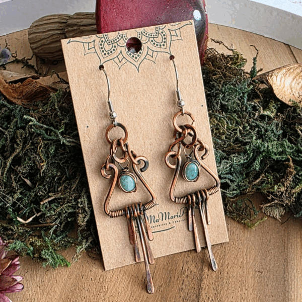 Handgefertigte Ohrringe aus geschwungenem Kupferdraht mit patinierter Oberfläche, verziert mit einem grünen Amazonit-Stein in der Mitte. Mehrere kleine Kupferstäbchen hängen beweglich an der Unterseite. Die Ohrringe sind auf einer Schmuckkarte mit Mandala-Druck arrangiert und liegen in einem naturinspirierten Set aus Moos, Trockenblumen und einem dekorativen roten Pilz.