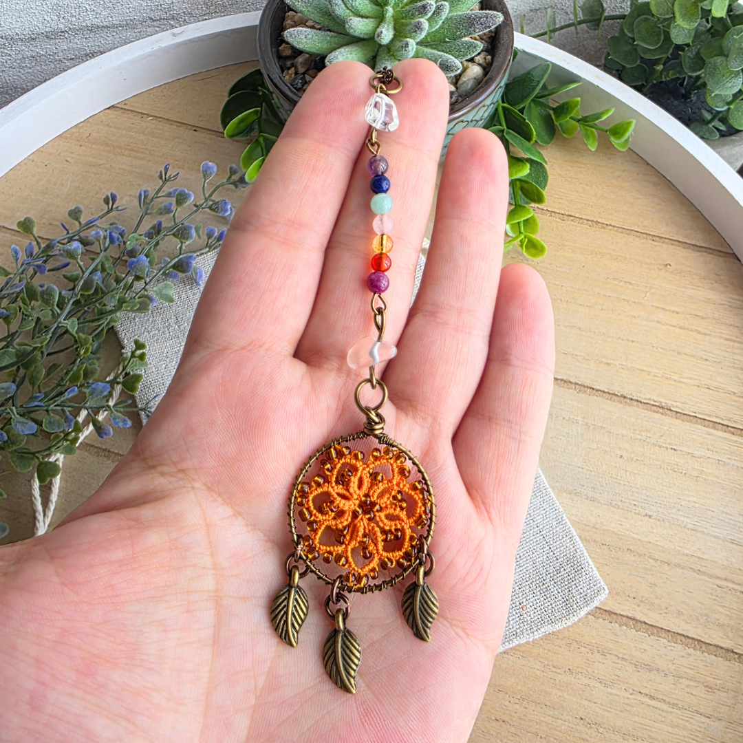 Größenansicht des handgefertigten Autospiegelanhängers mit orangefarbenem Occhi-Traumfänger und sieben Chakra-Edelsteinen in der Hand.